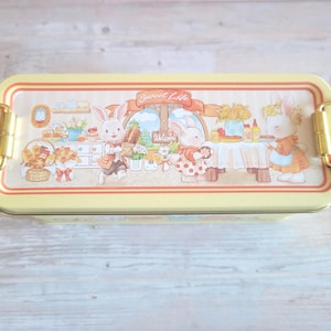 May include: A rectangular yellow metal lunch box featuring a whimsical illustration of three rabbits in a bakery setting with the text Sweet Life. The container has gold-toned metal latches on both sides and a light-colored border design.