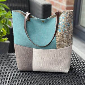 Peut inclure: Un sac fourre-tout patchwork avec un dessus turquoise, un fond marron et beige, et un côté à motifs. Le sac a des poignées en cuir marron et une fermeture à bouton. Le sac est fait de tissu texturé.