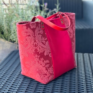 Peut inclure: Un sac cabas rose avec un motif floral d'un côté et rose uni de l'autre. Le sac a des anses en cuir et une fermeture éclair.