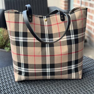 Peut inclure: Un grand sac fourre-tout à motif écossais beige, noir, rouge et blanc. Le sac est doté de poignées en cuir noir et d'une fermeture à bouton-pression dorée. Il est posé sur une surface métallique grise foncée.