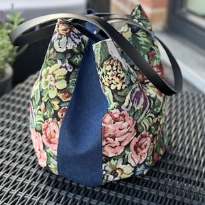 Op de afbeelding: Een tote bag van bloemenbehang met zwarte leren handvatten en een donkerblauw denim paneel. De tas heeft een grote, open bovenkant en een ruim interieur.