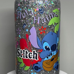 May include: A clear tumbler filled with silver glitter and colorful accents. The tumbler features a cartoon image of Stitch playing a ukulele, with the words "Hula Harmony" and "Stitch" in a playful font. Floral and tropical leaf designs are also present.