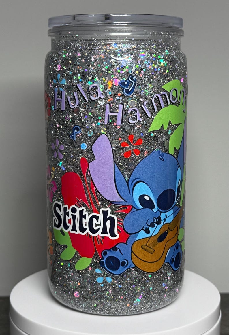 May include: A clear tumbler filled with silver glitter and colorful accents. The tumbler features a cartoon image of Stitch playing a ukulele, with the words "Hula Harmony" and "Stitch" in a playful font. Floral and tropical leaf designs are also present.