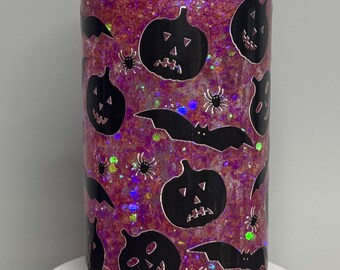 May include: A clear, cylindrical tumbler filled with pink glitter and decorated with black Halloween-themed cutouts. The design includes jack-o'-lanterns, bats, and spiders. The tumbler sits on a white surface.