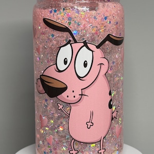 May include: A pink tumbler with a pink lid filled with pink glitter and a cartoon image of Courage the Cowardly Dog. The tumbler has a pink heart design and is on a white surface.