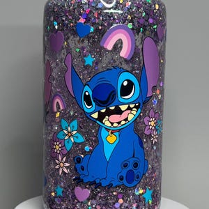 May include: A clear tumbler filled with purple glitter and featuring a blue cartoon character, Stitch, with large ears and a wide grin. The design includes hearts, stars, and flowers. The tumbler has a clear lid.