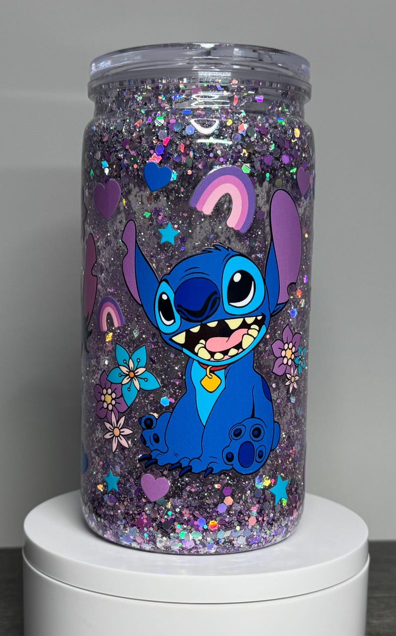 May include: A clear tumbler filled with purple glitter and featuring a blue cartoon character, Stitch, with large ears and a wide grin. The design includes hearts, stars, and flowers. The tumbler has a clear lid.