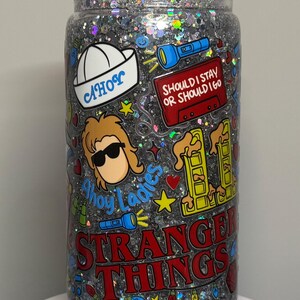 May include: Glitter-filled tumbler with a clear lid, adorned with colorful stickers. The stickers feature the text "Stranger Things", "Should I Stay or Should I Go", and "Ahoy Ladies", along with other themed graphics. The tumbler is filled with silver glitter.