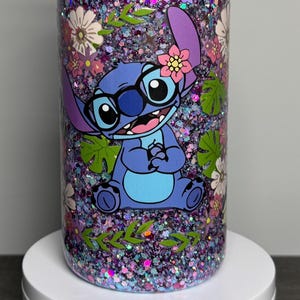 May include: A glitter-filled tumbler featuring a cartoon character with glasses and a flower. The tumbler has a floral and leaf design with a mix of purple, pink, and green glitter. The character is blue with a pink flower.