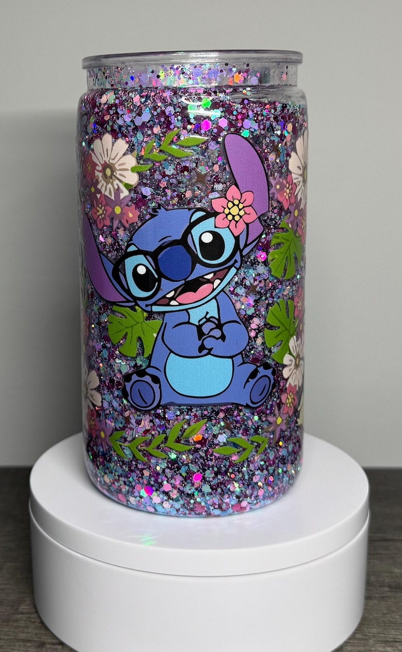 May include: A glitter-filled tumbler featuring a cartoon character with glasses and a flower. The tumbler has a floral and leaf design with a mix of purple, pink, and green glitter. The character is blue with a pink flower.