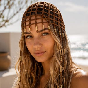 May include: A brown crocheted head covering with a grid pattern and dangling gold-colored beads. The headpiece is worn with long, wavy blonde hair. The image is taken outdoors with a blurred background of the ocean and sky.