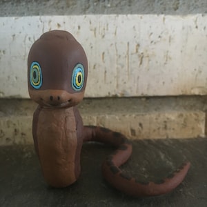 May include: A brown clay snake figurine with a curled tail. The snake has painted blue eyes with yellow rings around them.