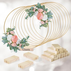 Könnte beinhalten: Ein goldfarbener Metallring-Hintergrund mit floralen Arrangements aus rosa und weißen Blumen und grünen Blättern. Der Text "12sets" befindet sich auf einem Holzblock. Mehrere Holzblöcke sind ebenfalls im Bild, geeignet für Eventdekorationen.