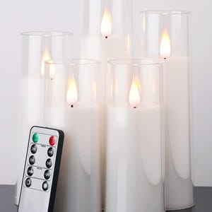 Op de afbeelding: Een set van vijf witte LED-pijlerkaarsen in heldere glazen cilinders. Elke kaars heeft een flikkerend vlameffect. Een afstandsbediening met diverse instellingen is inbegrepen.
