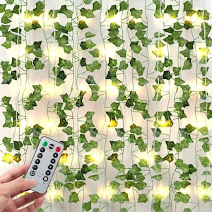May include: Green artificial ivy vines with small, warm white LED lights. A remote control is in the foreground. The vines are hanging, creating a decorative backdrop. The leaves are a mix of light and dark green.