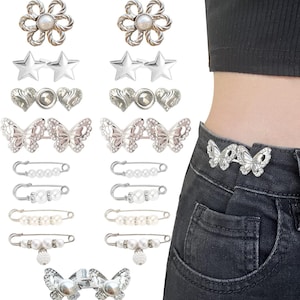 May include: A set of 12 silver-toned metal and pearl embellishments for clothing. The set includes two flower shapes, two star shapes, two heart shapes, two butterfly shapes, and four safety pins with pearl accents. The embellishments are shown on a pair of jeans.