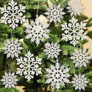 Op de afbeelding: Witte glitter sneeuwvlok ornamenten hangen aan een groene kerstboom. De sneeuwvlokken hebben ingewikkelde ontwerpen en zijn opgehangen aan dunne witte touwtjes. De naalden van de boom zorgen voor een contrasterende achtergrond.