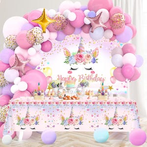 Op de afbeelding: Een verjaardagsfeestje met eenhoornthema. De achtergrond toont een eenhoorngezicht en de tekst "Happy Birthday". Een boog van roze, witte en gouden ballonnen. Een tafel is gedekt met een eenhoorn tafelkleed en taart.