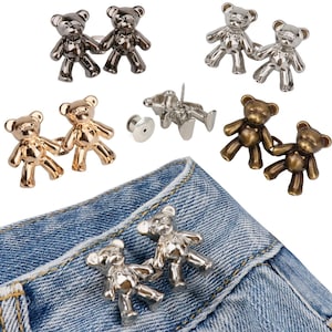Puede incluir: Surtido de botones metálicos en forma de osito de peluche en plata, oro y bronce. Los botones se muestran en un par de pantalones vaqueros azules.