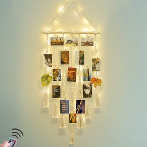 Puede incluir: Un expositor de fotos de macramé con luces de hadas. El colgante de pared presenta una clavija de madera, hilos de luces y múltiples cuerdas con pequeños clips de madera que sostienen varias fotos. Borlas decorativas y hojas añaden al diseño.