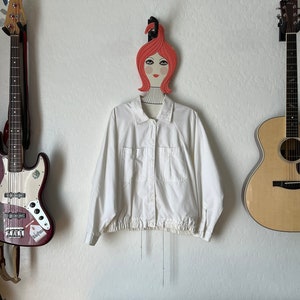 May include: A white jacket with a button-up front and two chest pockets hangs on a decorative wooden hanger shaped like a woman's face. A bass guitar and acoustic guitar are mounted on the wall on either side of the jacket.