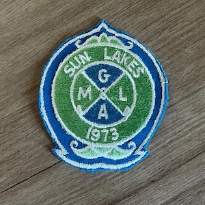 May include: Embroidered patch with a blue border and a green and white center. The patch reads "SUN LAKES" and "M & L G A 1973" in white lettering. The patch has a decorative top and bottom edge.