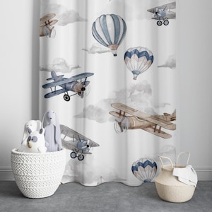 Könnte beinhalten: Weißer Vorhang mit einem sich wiederholenden Muster aus blauen und weißen Heißluftballons und braunen Flugzeugen. Der Vorhang hängt in einem Raum mit einem weißen Weidenkorb und zwei Stofftieren.