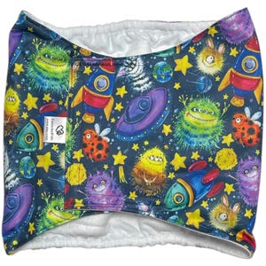 May include: A dark blue baby diaper cover with a colorful space-themed pattern. The design includes rockets, planets, stars, and cartoon aliens. The interior is white.