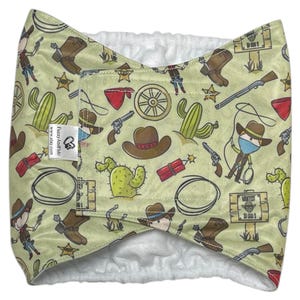 May include: A light green fabric item with a repeating pattern of cowboy boots, hats, cacti, revolvers, and cartoon cowboys. The item has a white, fluffy interior and a tag with text.