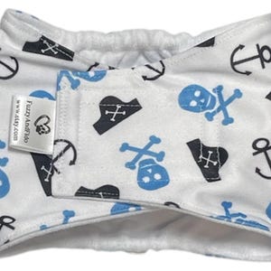 May include: White pet belly band with a pirate theme. The fabric features blue and black skull and crossbones, anchors, and pirate hat patterns. A small tag reads "FuzzyAndFido" and "www.etsy.com".