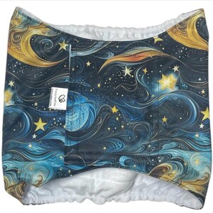 May include: A reusable cloth diaper featuring a celestial design with swirling blues, golds, and white. The pattern includes stars and crescent moons. The diaper has a white interior and a small tag with text.