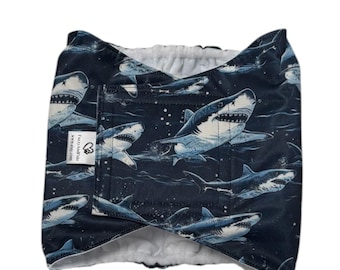 May include: A dark blue cloth diaper with a repeating pattern of white and blue sharks swimming in the ocean. The diaper has a white interior and a tag with text and a heart logo.