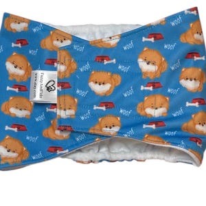 May include: Blue dog-themed pet accessory with a repeating pattern of orange Pomeranian illustrations, dog bowls, and bones. The word "woof" is also printed on the fabric. The tag reads "FuzzyAndFido" and "www.etsy.com".