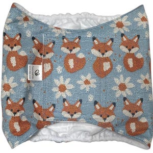 May include: A blue cloth diaper cover featuring a repeating pattern of orange foxes and white daisies. The diaper has a white interior and a small tag with text.