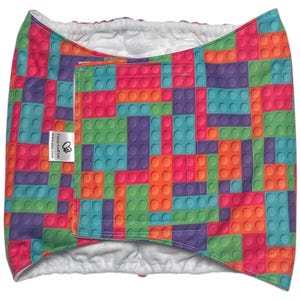 May include: A colorful, patterned wrap featuring a design of toy building blocks in shades of pink, orange, green, purple, and teal. The wrap has a white interior and a velcro closure. A small tag with text is visible.