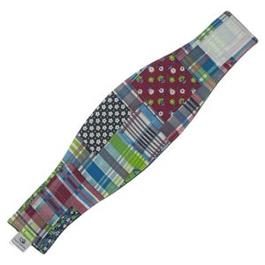 May include: A patchwork fabric wrap in a curved shape, featuring a variety of patterned squares in shades of blue, green, red, and white. The wrap has a white velcro closure and a small tag with text.