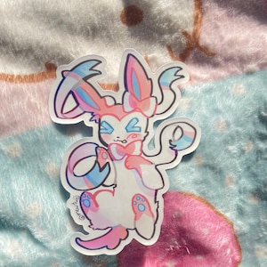 Puede incluir: Una pegatina transparente de un personaje Pokémon rosa y azul con un cuerpo blanco y ojos azules. El personaje sostiene una cinta rosa y azul en su mano derecha y un círculo rosa y azul en su mano izquierda. La pegatina tiene un acabado holográfico y está sobre un fondo azul y rosa.