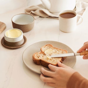 May include: A person spreading butter on a slice of bread with a butter knife. The bread is on a beige plate. There is a small dish of butter and a mug of tea on the table.