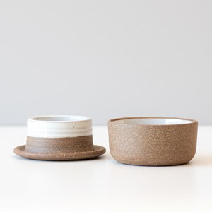 May include: A set of two ceramic butter dishes. The smaller dish has a white glaze on the top half and a brown speckled glaze on the bottom half. The larger dish is entirely brown speckled.