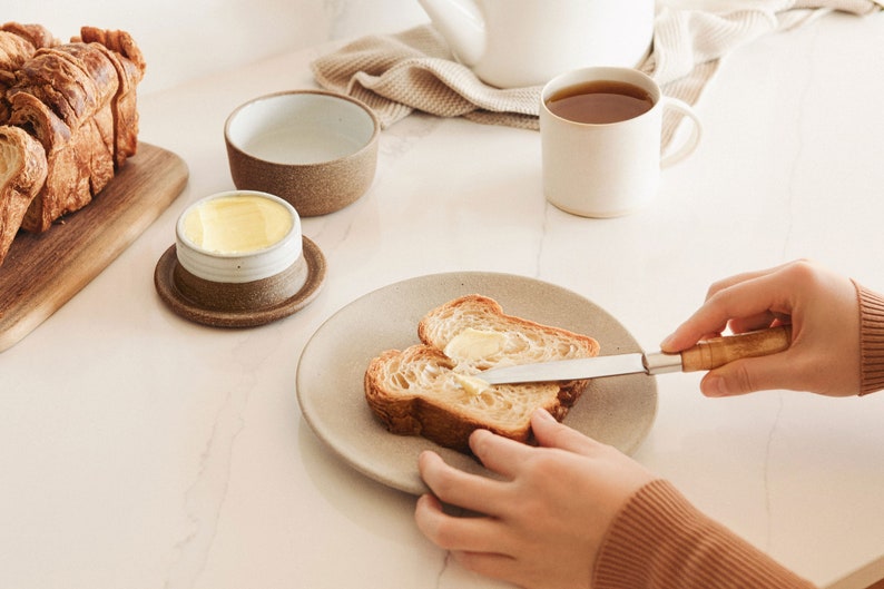 Pode incluir: Uma pessoa espalhando manteiga em uma fatia de p&atilde;o com uma faca de manteiga. O p&atilde;o est&aacute; em um prato bege. H&aacute; um pequeno pote de manteiga de cer&acirc;mica branca com uma borda marrom em um pires marrom. H&aacute; uma caneca de cer&acirc;mica branca com uma borda marrom cheia de ch&aacute;.
