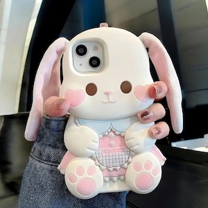 Puede incluir: Funda de teléfono con forma de conejo blanco y rosa, con orejas grandes, mejillas en forma de corazón y pies con forma de pata. La funda tiene un vestido rosa y el texto "rabbiteleven" es visible. Diseñado para smartphones.