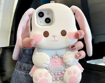 May include: A white and pink bunny phone case with large ears, heart-shaped cheeks, and paw-shaped feet. The case has a pink dress and the text "rabbiteleven" is visible on the front. The phone case is designed to fit a smartphone.