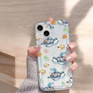 Puede incluir: Carcasa transparente para teléfono con un diseño de tortuga marina. La carcasa tiene un fondo azul claro con detalles en blanco y amarillo. Las tortugas están rodeadas de algas marinas y conchas.