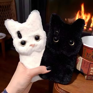 May include: Two fluffy cat-shaped phone cases, one white with a black spot and pink nose, the other black. Both have large, round eyes and are held together. The cases are designed to resemble a cat's face.