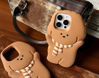 Puede incluir: Dos fundas de teléfono marrones con forma de oso de dibujos animados con una bufanda a rayas. Las fundas tienen una cara sonriente y brazos. Un bolso marrón y beige está en el fondo.