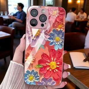 May include: A phone case with a floral design in red, blue, and purple, with yellow centres. The case has a clear, glossy finish and a raised, textured appearance. The phone case has a pink border and a camera module.