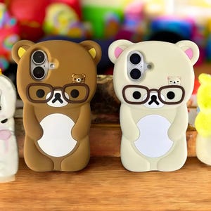May include: Two phone cases shaped like cartoon bears. One is brown, the other is cream-colored. Both have black-rimmed glasses and small bear faces. The cases have cutouts for the phone's camera and buttons. A small bear face is also on the top of each case.