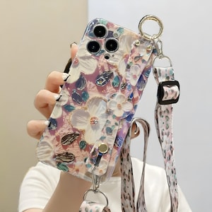 Könnte beinhalten: Eine transparente Handyhülle mit floralem Design. Die Hülle zeigt weiße, rosa und blaue Blumen mit goldenen Akzenten. Sie enthält einen passenden Gurt mit floralem Muster und einen goldfarbenen Metallring zur Befestigung.