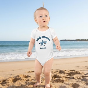 Könnte beinhalten: Ein weißer Baby-Body mit einem blau-weißen Hai-Motiv und dem Text "Totally Jawsome CAPE COD". Das Baby steht an einem Sandstrand mit dem Meer im Hintergrund.