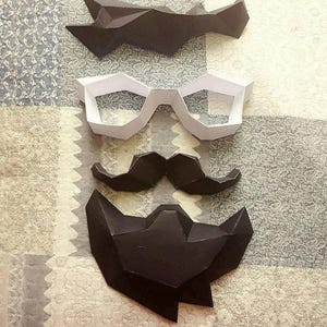 May include: A set of three papercraft masks, including a black mustache, black beard, and white glasses. The masks are made of folded paper and have a geometric design.